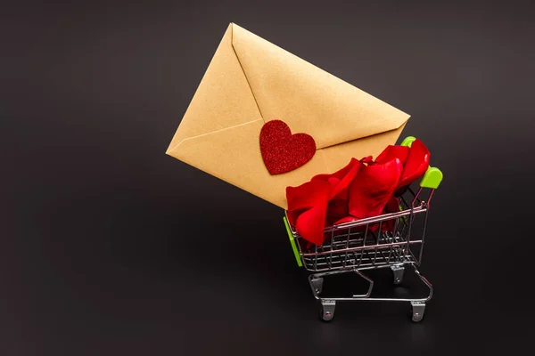 Shopping cart with rose petals and envelope isolated on black — Stock Photo