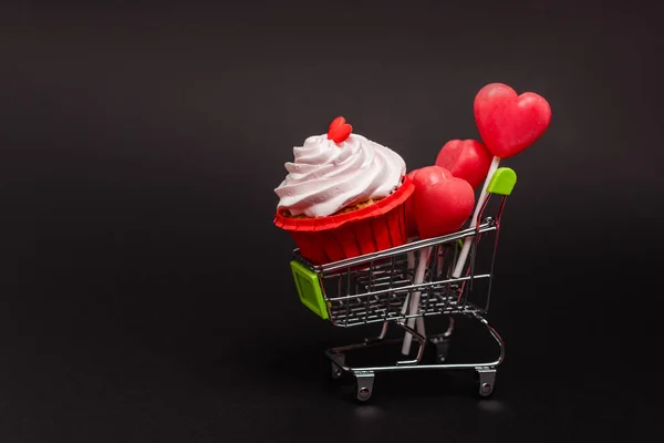 Shopping cart with valentines cupcake and lollipops isolated on black — Stock Photo