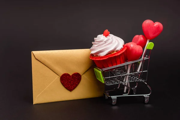 Carrito de compras con cupcake de San Valentín y piruletas y sobre aislado en negro - foto de stock