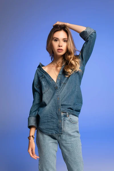 Young, charming woman in denim shirt and jeans posing with raised hand isolated on blue — Stock Photo