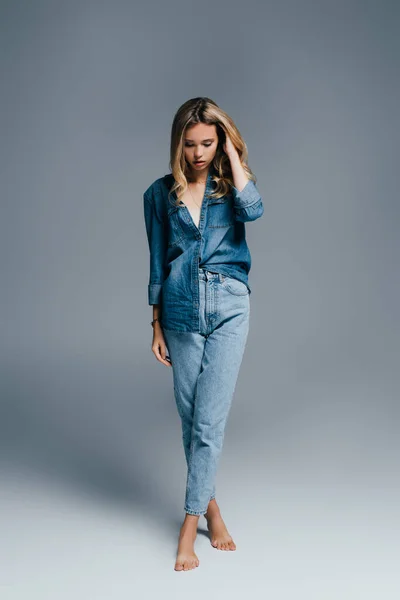 Young woman in denim shirt and jeans touching hair while standing barefoot on grey — Stock Photo