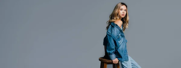 Young woman in denim shirt, with naked shoulder, leaning on high chair isolated on grey, banner — Stock Photo