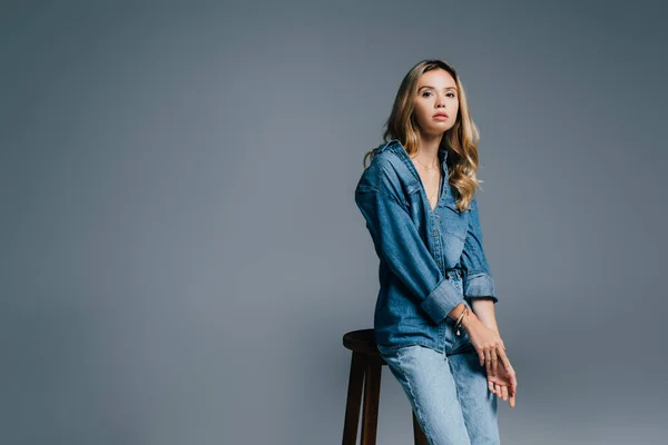 Mujer con estilo en ropa de mezclilla cerca de taburete alto aislado en gris - foto de stock