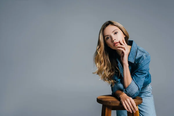 Sensual young woman in denim shirt looking at camera while leaning on high stool isolated on grey — Stock Photo