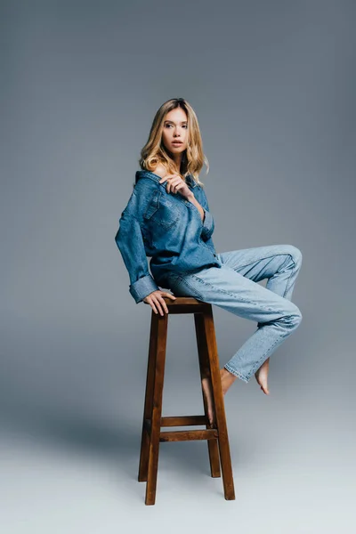 Seductive barefoot woman in denim clothes, with naked shoulder, sitting on high chair on grey — Stock Photo