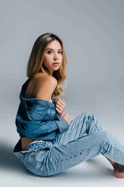 Sexy young woman in unbuttoned denim shirt and jeans posing with naked shoulder on grey — Stock Photo