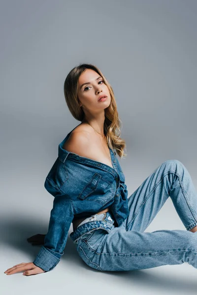 Seductora joven en jeans desabotonados, con hombro desnudo, sentada sobre gris - foto de stock