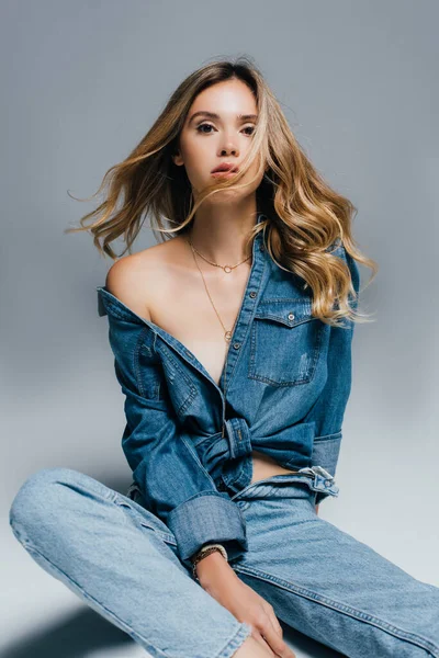 Sexy joven mujer en desabotonado denim camisa y jeans mirando a la cámara mientras posando en gris - foto de stock