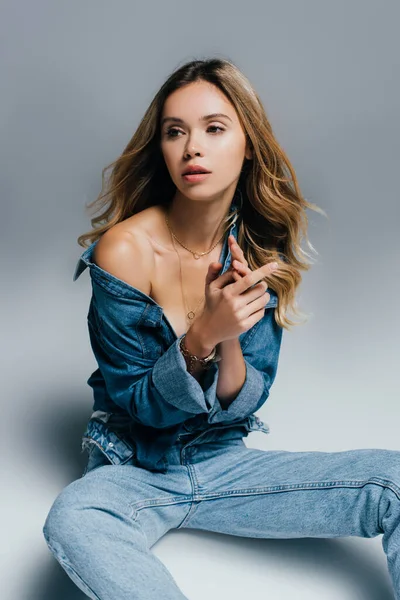 Sexy jeune femme en denim déboutonné chemise regardant loin tout en étant assis sur gris — Photo de stock