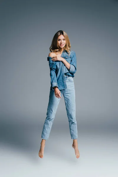 Barefoot woman in denim clothes, with naked shoulder, smiling at camera while levitating on grey — Stock Photo
