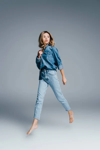 Молодая босиком женщина в джинсовой рубашке и джинсах смотрит в сторону, левитируя на сером — стоковое фото