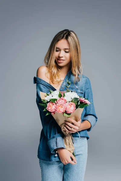 Charming woman in denim shirt, with naked shoulder, posing with bouquet isolated on grey — Stock Photo
