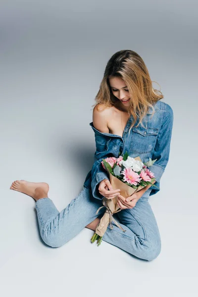 Високий кут зору усміхненої жінки з голим плечем, одягнений в джинсовий одяг, сидить з квітами на сірому — стокове фото