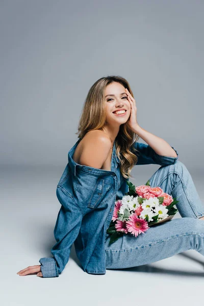 Allegra giovane donna vestita in denim, con spalla nuda, seduta con bouquet su grigio — Foto stock