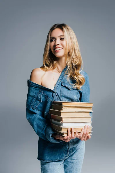 Молода спокуслива жінка в джинсовій сорочці, з голим плечем, тримає книги ізольовані на сірому — стокове фото