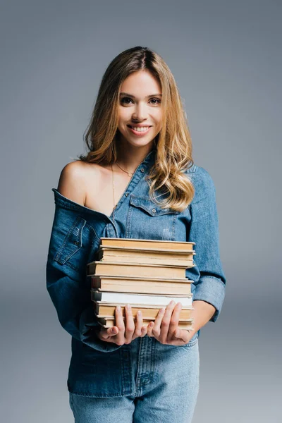 Young smiling woman in denim shirt holding stack of books while standing with naked shoulder isolated on grey — Stock Photo