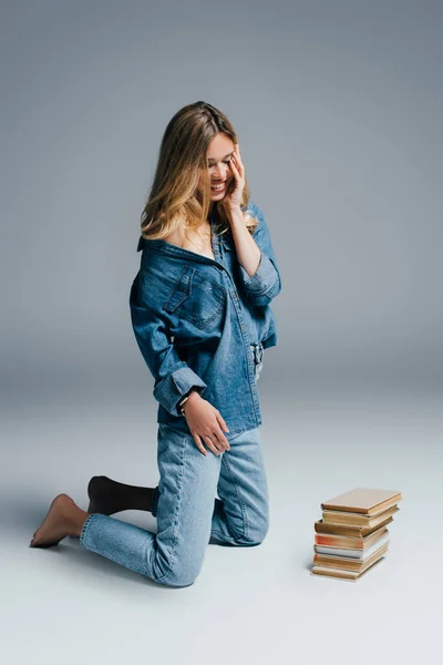 Donna sorridente in abiti di denim toccare il viso mentre si inginocchia vicino a libri sul grigio — Foto stock