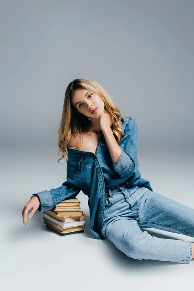 Seductive woman in denim shirt and jeans, with naked shoulder, leaning on books while sitting on grey — Stock Photo