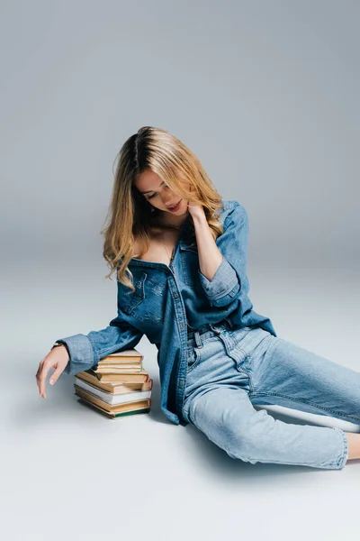 Молодая соблазнительная женщина в джинсовой одежде трогает шею и опирается на книги, сидя на сером — стоковое фото