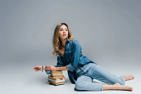 Joven mujer seductora en ropa de mezclilla mirando hacia otro lado mientras se apoya en libros sobre gris - foto de stock