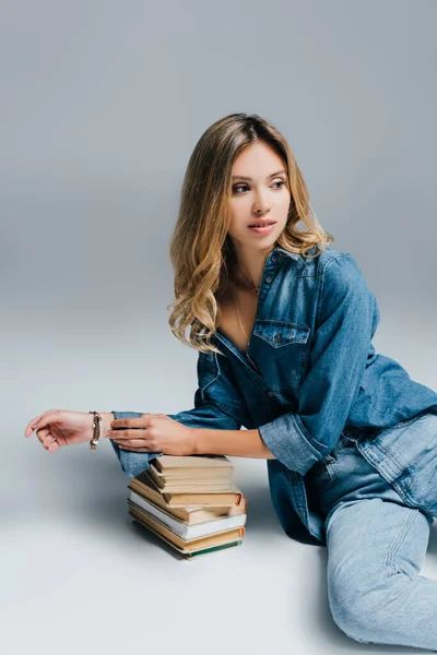 Чарівна жінка в джинсовій сорочці і джинсах спирається на книги, сидячи на сірому — стокове фото