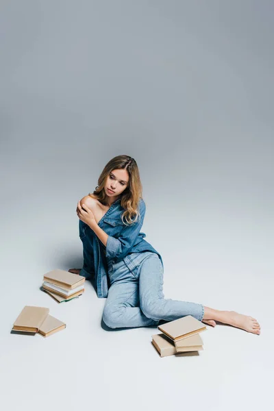 Giovane donna sensuale in abiti di denim toccare spalla nuda mentre seduto vicino a libri sul grigio — Foto stock