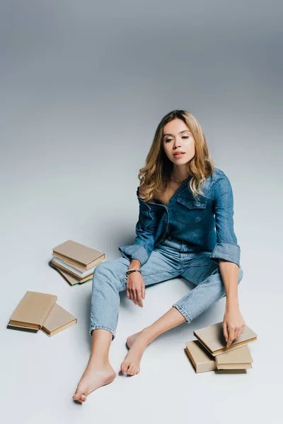 Young barefoot woman in denim shirt and jeans sitting near books on grey — Stock Photo