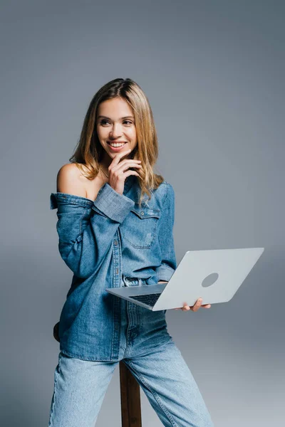Улыбающаяся женщина в джинсовой рубашке и джинсах смотрит в сторону, держа ноутбук на сером — стоковое фото