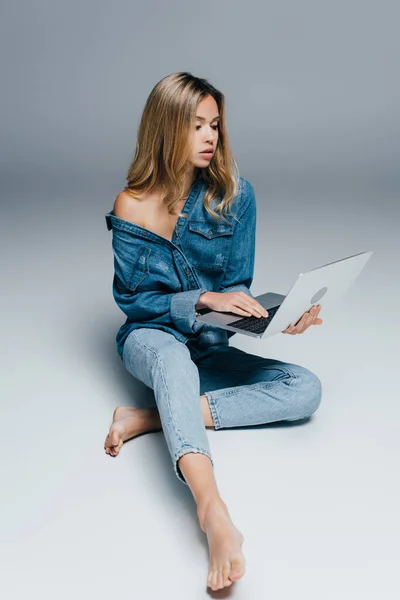 Sensuelle pieds nus femme en vêtements en denim, avec épaule nue, en utilisant un ordinateur portable tout en étant assis sur gris — Photo de stock