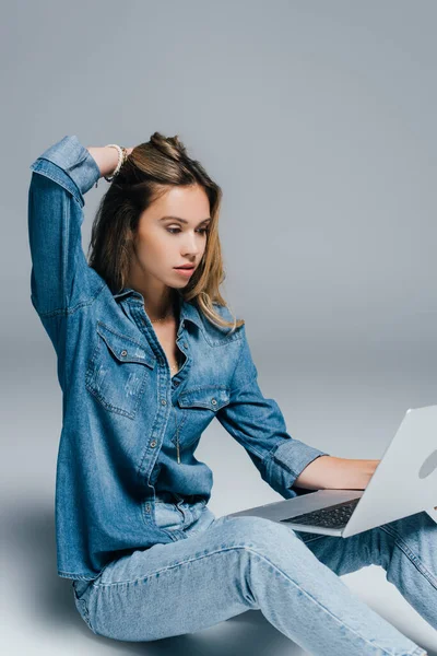 Jeune, élégant femme en denim vêtements toucher les cheveux tout en étant assis avec ordinateur portable sur gris — Photo de stock