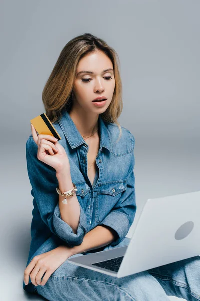 Femme élégante en vêtements denim tenant carte de crédit tout en regardant ordinateur portable sur gris — Photo de stock