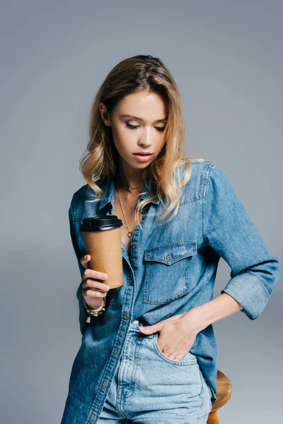 Sensual jovem mulher em camisa jeans e jeans, com a mão no bolso, segurando café para ir isolado em cinza — Fotografia de Stock