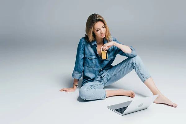 Barefoot woman in denim shirt and jeans holding credit card while sitting on grey near laptop — Stock Photo