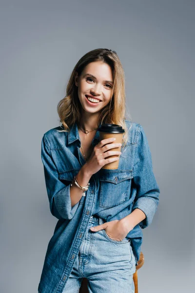 Улыбающаяся молодая женщина в джинсовой одежде держа руку в кармане и кофе, чтобы пойти изолированы на серый — стоковое фото