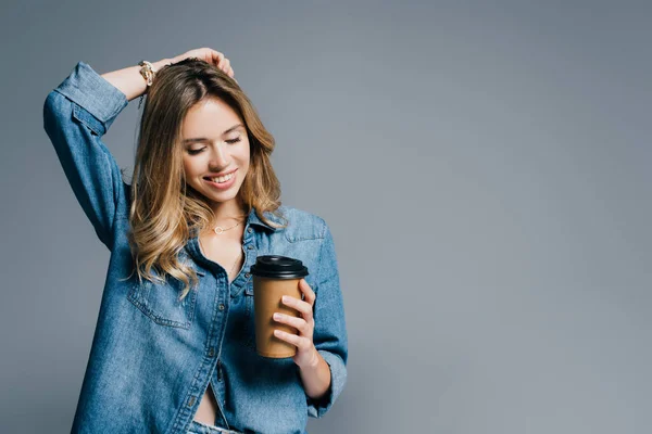 Femme souriante en denim toucher les cheveux tout en tenant du café pour aller isolé sur gris — Photo de stock