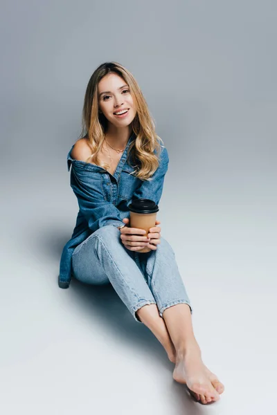 Barefoot woman wearing denim clothes, with naked shoulder, sitting with paper cup on grey background — Stock Photo