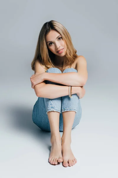 Sensual barefoot woman in jeans, with naked shoulders, looking at camera while sitting on grey — Stock Photo