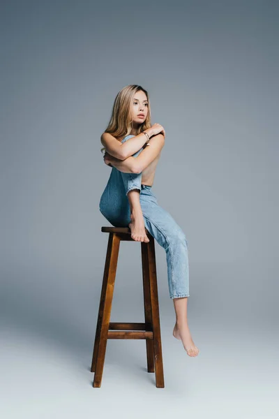 Соблазнительная топлесс женщина в джинсах смотрит в сторону, сидя на стульчике на сером — стоковое фото