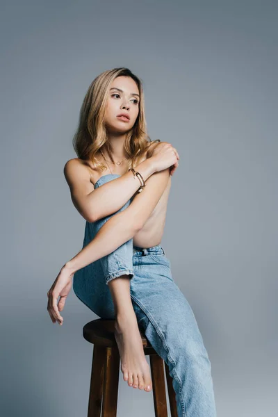 Topless, barefoot woman in jeans sitting on high stool isolated on grey — Stock Photo