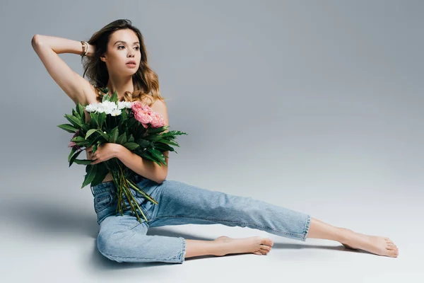 Sexy, donna a piedi nudi in jeans, con le spalle nude, toccando i capelli mentre seduto con i fiori sul grigio — Foto stock