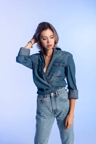 Mujer joven y seductora en ropa de mezclilla con estilo tocando el cabello aislado en azul - foto de stock