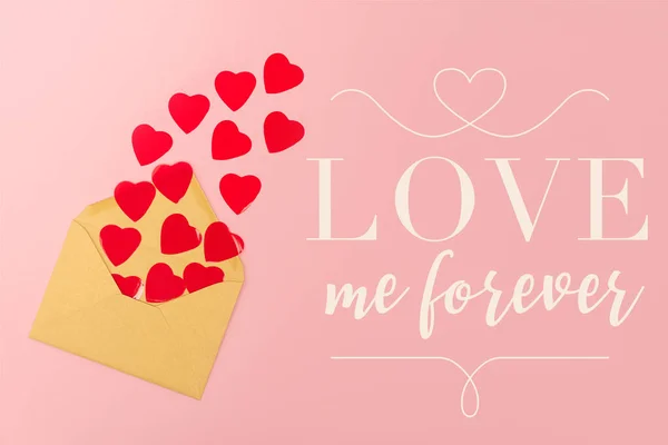 Top view of red paper hearts and envelope near love me forever lettering on pink background — Stock Photo