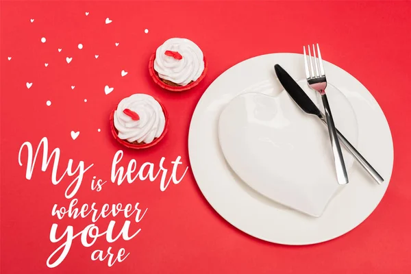 Top view of cupcakes near plates with cutlery and my heart is wherever you are lettering on red background — Stock Photo