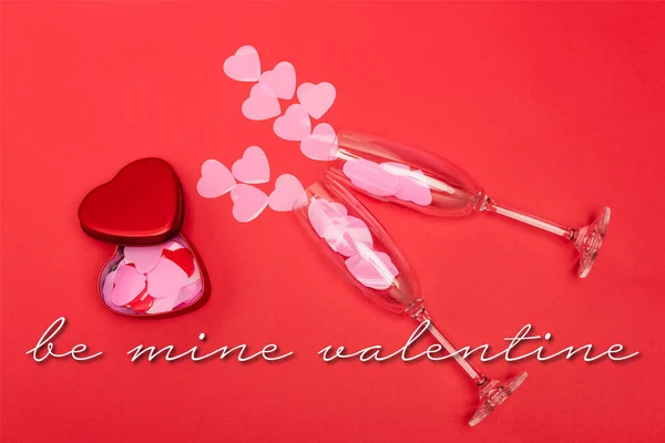 Top view of pink hearts in champagne glasses near metallic box and be mine valentine lettering on red background — Stock Photo