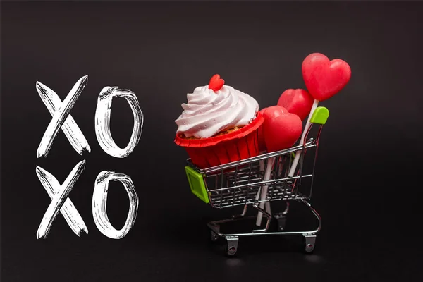 Chariot d'achat de jouets avec des valentines, cupcake et sucettes près de lettrage xoxo sur noir — Photo de stock