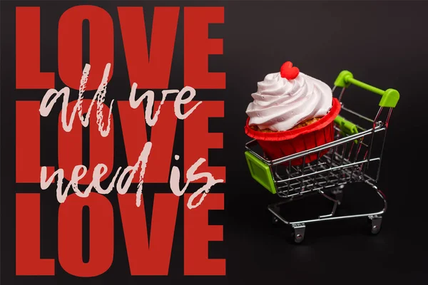 Pequeño carrito de compras con cupcake de San Valentín cerca de todo lo que necesitamos es letras de amor en negro - foto de stock