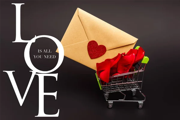 Toy shopping cart with rose petals and envelope near love is all you need lettering on black — Stock Photo