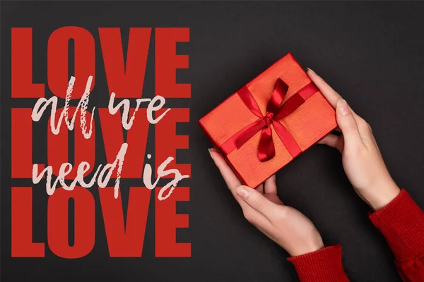 Cropped view of woman holding red gift near all we need is love lettering on black — Stock Photo