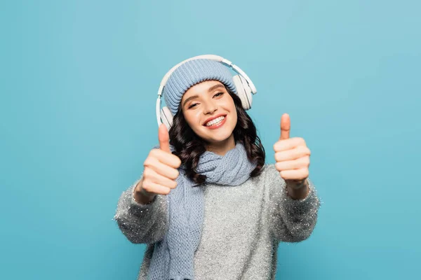 Happy woman in hat and scarf listening to music in headphones and showing thumbs up isolated on blue — Stock Photo