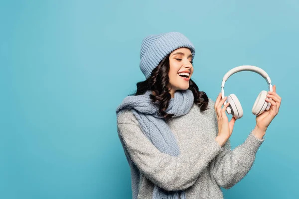 Felice donna bruna in abito invernale guardando le cuffie isolate sul blu — Foto stock